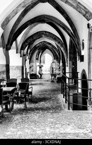 Schwarzweiß-Foto von Menschen, die durch gotische Gewölbearkade mit Café-Sitzplätzen in Prag, Tschechische Republik, spazieren. Stockfoto