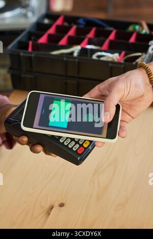 Kaukasischer junger Erwachsener, der ein Smartphone über dem Zahlungsterminal hält und kontaktlos bezahlt, während Schwarze Frau mit Kartenleser an der Kasse eines Second-Shop hält Stockfoto