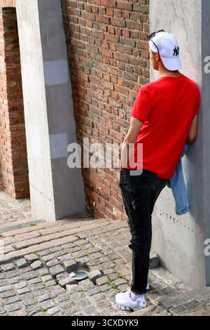 Junger Mann in rotem T-Shirt und Mütze, der sich an eine Säule in einer städtischen Umgebung lehnt Stockfoto