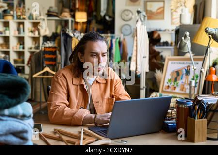 Kaukasischer junger Erwachsener sitzt am Schreibtisch mit Laptop, umgeben von Kleiderständern und Gebrauchtgegenständen in einem Second-Hand-Shop, wobei er sich während der Arbeit oder der Bestandsverwaltung auf den Bildschirm konzentriert Stockfoto
