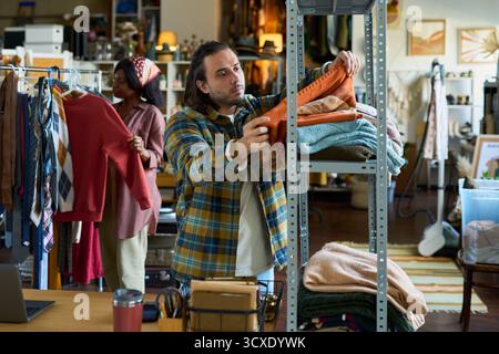 Kaukasier mittleren Alters organisierte gefaltete Kleidung auf Metallregalen, während Schwarze in Second-Hand-Geschäften hängende Kleidung stöberte. Beide konzentrierten sich auf das Sortieren und Auswählen von Second-Hand-Kleidungsstücken Stockfoto
