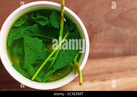 Frische Minze in einer Tasse heißem Wasser auf Holztisch. Heilende beruhigende Kräuterteebrühung Stockfoto
