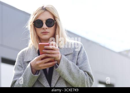 Porträt der modischen Transfrau. Flacher Blick auf blonden Transgender in runder Sonnenbrille und stilvollem Mantel mit Smartphone und SMS Stockfoto