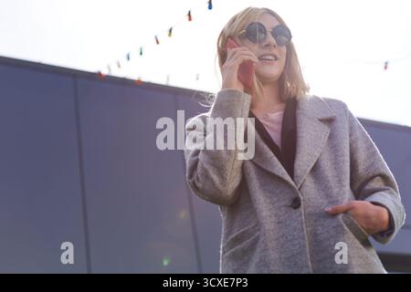 Porträt einer androgynen Geschäftsfrau. Flacher Blickwinkel einer selbstbewussten, stilvollen blonden Transfrau in Sonnenbrille, die auf dem Smartphone spricht und lächelt Stockfoto