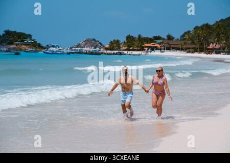 Ein Mann und eine Frau rennen zusammen am Strand, halten Hände und lächeln. Die helle Sonne scheint herunter, während sanfte Wellen zu ihren Füßen schlagen und einen erzeugen Stockfoto