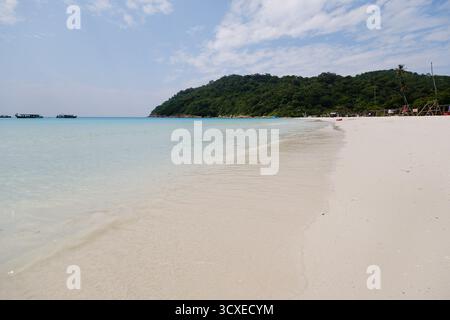 Die Wellen schlagen sanft gegen die Küste eines Sandstrandes, umgeben von üppigen grünen Hügeln. Das klare türkisfarbene Wasser glitzert unter dem Sonnenlicht und erzeugt einen Ser Stockfoto