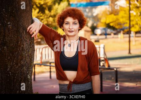 Porträt einer erwachsenen kaukasischen Frau mit lockigen Haaren, die in der Nähe eines Baumes auf dem Hintergrund des Sportplatzes posiert. Stockfoto