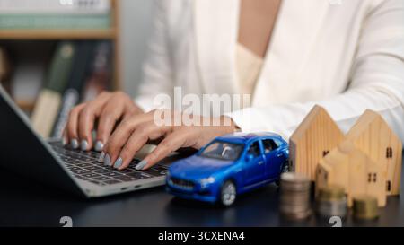 Geschäftsfrau, die auf einem Laptop mit Automodell, Hausmodell und Münzstapel auf dem Schreibtisch schreibt, die Finanzplanung, Immobilieninvestitionen und c Stockfoto