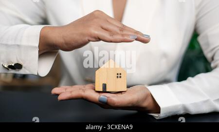 Geschäftsfrau, die mit ihren Händen ein hölzernes Hausmodell schützt, symbolisiert Hausversicherung, Sachschutz, Immobilieninvestitionen und Finanzen Stockfoto