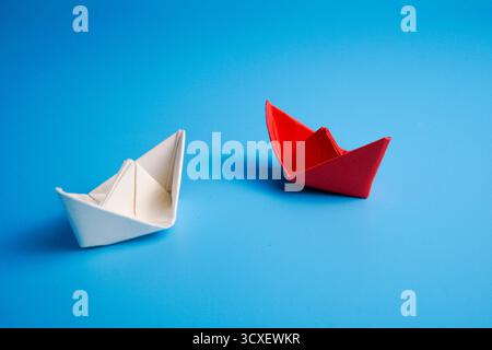 Minimalistisches Konzept mit zwei Papierbooten, eines rot und eines weiß, die sich auf blauem Hintergrund gegenüberstehen. Symbolisieren Führung, Kommunikation. Stockfoto