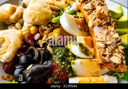 Nahaufnahme von geröstetem Lachssteak mit Avocado, Müsli, gekochten Eiern, Oliven und gemischtem frischem Gemüse auf weißer Keramikplatte auf einem Tisch. Stockfoto