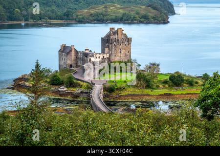 Eilean Donan Castle befindet sich auf einer winzigen Insel, die durch eine gekrümmte Steinbrücke verbunden ist. Eine schottische Flagge fliegt neben gepflegten Grundstücken als ruhiges Wasser und weit entfernt Stockfoto