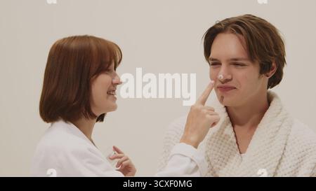 Fröhliche Frau im weißen Bademantel, die Hautpflegecreme auf die lächelnde Männernase aufträgt. Stockfoto