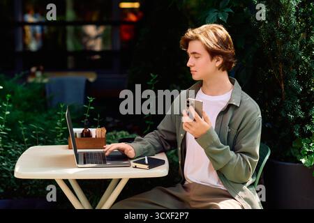 Ein fokussierter junger Mann arbeitet an einem Laptop in einem belebten Café, umgeben von Grün und modernem Flair. Stockfoto