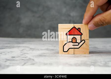 Holzwürfel mit Illustration eines auf der Hand ruhenden Hauses mit rotem Dach als Symbol für Sachversicherung, Wohnungsinvestition, Hypotheken oder Immobilienpflege. Stockfoto
