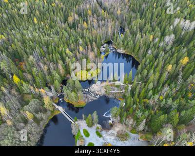 Blick aus einem hohen Winkel auf den üppigen grünen Wald und das gelbe Herbstlaub, das einen dunklen, still liegenden See mit einer Holzbrücke und kleiner touristischer Infrastruktur umgibt Stockfoto