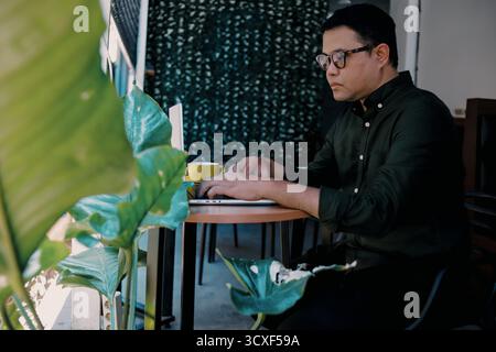 Junger asiatischer Mann, der mit Laptop im Coffee Shop arbeitet, während er Kaffee trinkt Stockfoto
