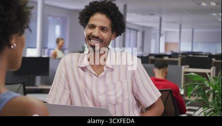 Lächelnder hispanischer Mann in gestreiftem Hemd, der mit einem Kollegen im Großraumbüro spricht, mit Laptop-Monitoren Stockfoto