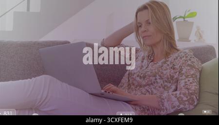 Eine Tippfrau mit Blumenbluse, die am Laptop auf dem Sofa im Wohnzimmer arbeitet, mit Pflanze, Figur Stockfoto