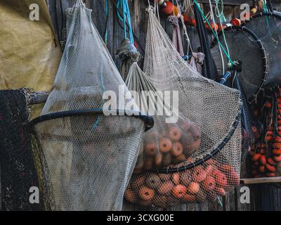 Traditionelle Fischernetze und orangene Schwimmer, die in Netztüten gelagert werden und an einer Holzwand im mediterranen Dorf hängen Stockfoto