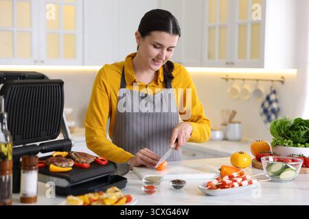 Frau, die Pfeffer schneidet, während sie Fleisch und Gemüse auf einem elektrischen Grill in der Küche kocht Stockfoto