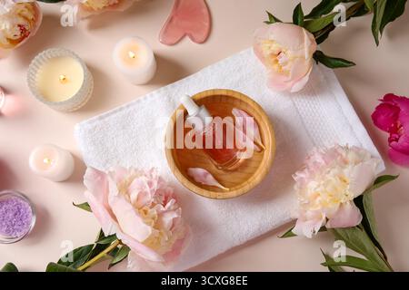 Flasche ätherisches Öl mit Kerzen, Meersalz, guasha-Massagewerkzeug und schöne Pfingstrosenblüten auf beigem Hintergrund Stockfoto