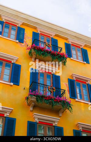 Farbenfrohe Fassade im mediterranen Stil mit blauen Fensterläden, roten Fensterrahmen und lebendigen Balkonen mit Blumendecken vor sonnigem Himmel Stockfoto