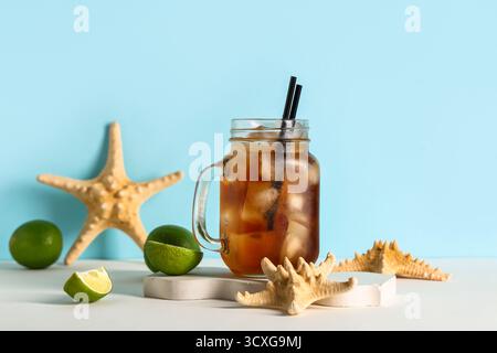 Mauerglas mit kaltem Cuba Libre Cocktail mit Sternenfischen und Gipspodium auf weißem Tisch vor blauem Hintergrund Stockfoto