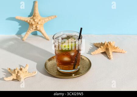 Glas kalter Cuba Libre Cocktail mit Sternenfischen auf weißem Tisch vor blauem Hintergrund Stockfoto