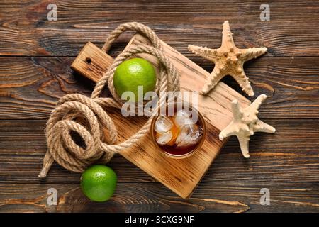 Bord mit einem Glas kalten Cuba Libre Cocktail, Sternenfischen und Seil auf hölzernem Hintergrund Stockfoto