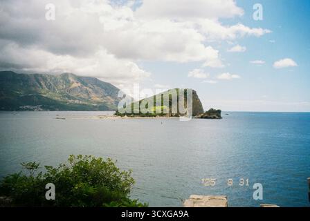 Malerischer Blick auf die Insel Sveti Nikola in der Nähe von Budva, Montenegro, mit üppigem Grün, felsigen Klippen und der Adria unter teilweise bewölktem Himmel auf Film Stockfoto