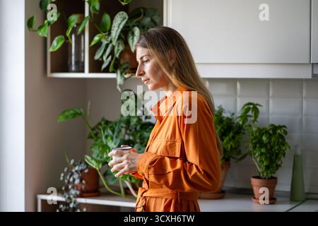 Einsame Frau mittleren Alters, die völlig in Gedanken verloren ist, mit einem warmen Teetasse, der in der Küche steht Stockfoto