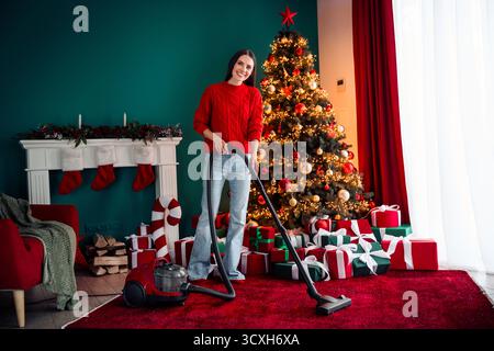 Charmante Frau saugt ein gemütliches Haus, das mit einem festlichen Weihnachtsbaum und Geschenken dekoriert ist und eine fröhliche Urlaubsatmosphäre schafft Stockfoto