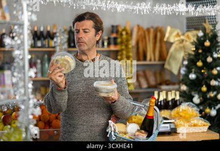 Konzentrierter erwachsener Mann mit Warenkorb, der versucht, eine Wahl zu treffen und zwei Heringsarten im Lebensmittelgeschäft zu halten, um sich auf die Weihnachtsfeier vorzubereiten Stockfoto