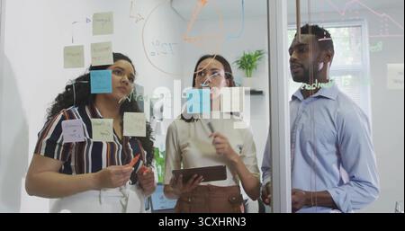 Brainstorming von Kollegen, die Haftnotizen auf Glastafel im Besprechungsraum mit Tablet und Markern untersuchen Stockfoto