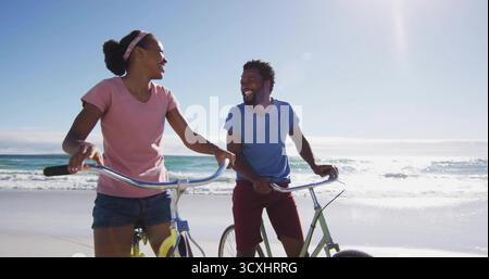 Lächelndes Paar, das unter Sonnenlicht an der Sandküste mit Fahrrädern unterwegs ist, T-Shirts und Shorts trägt Stockfoto