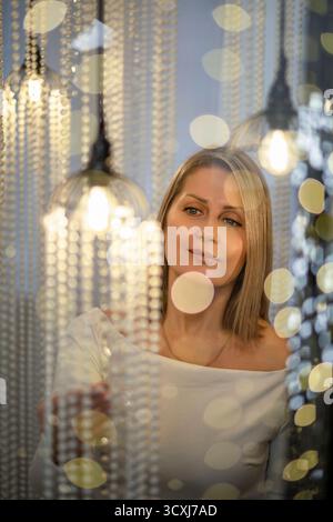 Eine junge kaukasische Frau mit blonden Haaren blickt nachdenklich inmitten glitzernder Weihnachtslichter. Die Szene vermittelt eine magische Urlaubsatmosphäre. Stockfoto
