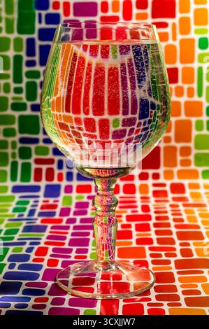 Klares Weinglas gefüllt mit spritziger Flüssigkeit, auf einer lebhaften Mosaikoberfläche platziert, wodurch sich brechende Farbverzerrungen ergeben. Stockfoto