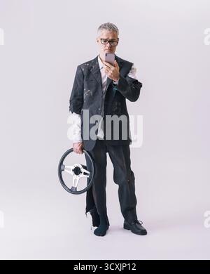 Lustiges Konzeptbild eines Mannes, der ein abgetrenntes Autolenkrad hält und auf sein Smartphone schaut, um Hilfe anzufordern. Symbol für Autounfall, mobile Insur Stockfoto