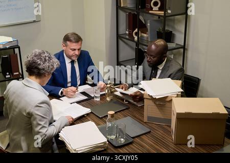 Kaukasischer Mann mittleren Alters und junger Schwarzer Mann sitzen mit einer älteren kaukasischen Frau am Tisch und prüfen Rechtsdokumente im Büro mit sichtbaren Akten und Papieren Stockfoto