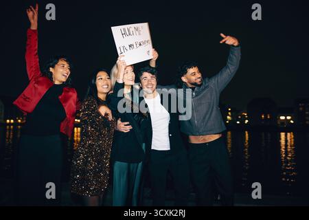 Eine fröhliche Gruppe von Freunden feiert nachts einen Geburtstag am Wasser. Lachen und Freude sind durchdrungen, wenn sie mit festlichen Outfits und Schildern posieren, cr Stockfoto
