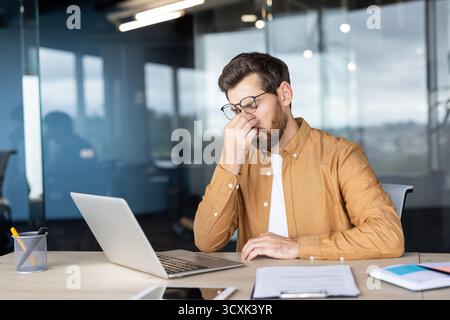 Geschäftsmann fühlt sich überfordert und müde, reibt sich die Augen vor Augenschmerzen und Kopfschmerzen, während er an einem modernen Schreibtisch an einem Laptop arbeitet, Symbolizi Stockfoto