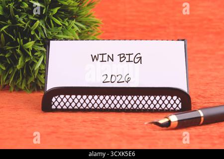 Eine Nachricht auf einem Schreibtisch besagt, dass WIN BIG 2026 neben einer dekorativen Pflanze und einem Stift auf einer strukturierten, orangefarbenen Oberfläche ein Zeichen für Planung und eine vielversprechende Zukunft ist. Stockfoto