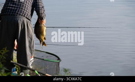 Mann, der am Flussufer steht und einen frisch gefangenen Fisch über dem Wasser hält, mit Angelruten im Hintergrund. Realistische Außenfotos für die Mo-Aufnahmen Stockfoto