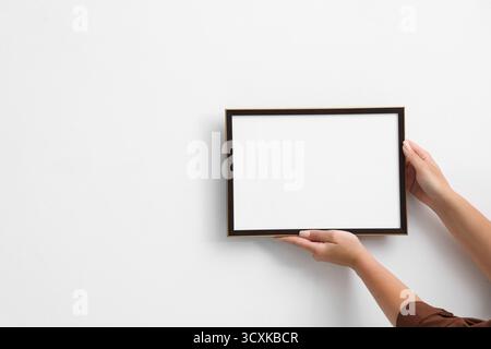 Frau, die einen leeren Diplomrahmen an der weißen Wand hängt, Großaufnahme. Modell für Design Stockfoto