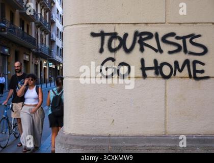 Die Leute gehen die Straße entlang, vorbei an einem Graffiti an einer Wand, auf dem steht "Touristen gehen nach Hause" in Barcelona, Spanien Stockfoto