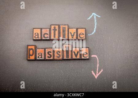 Aktiv und Passiv in hölzernen Buchstabenblöcken auf dunkler Oberfläche geschrieben, mit farbigen Kreidepfeilen, die nach oben zeigen, um aktiv zu sein, und nach unten, um passiv zu sein. Symbolisiert Lebensstil und persönliche Entwicklung. Stockfoto