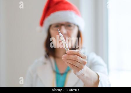 Nahaufnahme einer fröhlichen Ärztin in Sankt-Hut und medizinischer Uniform, lächelnd und mit Thermometer. Fürsorgliche Wintergrippe Check, Patientengesundheitsbewusstsein, Weihnachtszeit in der Krankenhausversorgung. Stockfoto