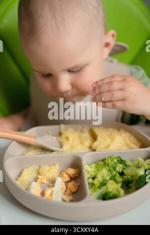 Happy Baby, das nahrhafte Essen mit den Händen erkundet – Maisbrei, gedämpfter Brokkoli und gekochte Eierstücke. Natürlicher Absetzprozess und Nahrungsentdeckungsmoment. Stockfoto