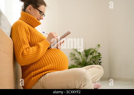 Heitere schwangere Frau schreibt wichtige Liefererinnerungen zu Hause im Notizbuch. Durchdachte Vorbereitung auf die Grundlagen der Entbindungs- und Neugeborenenpflege Stockfoto
